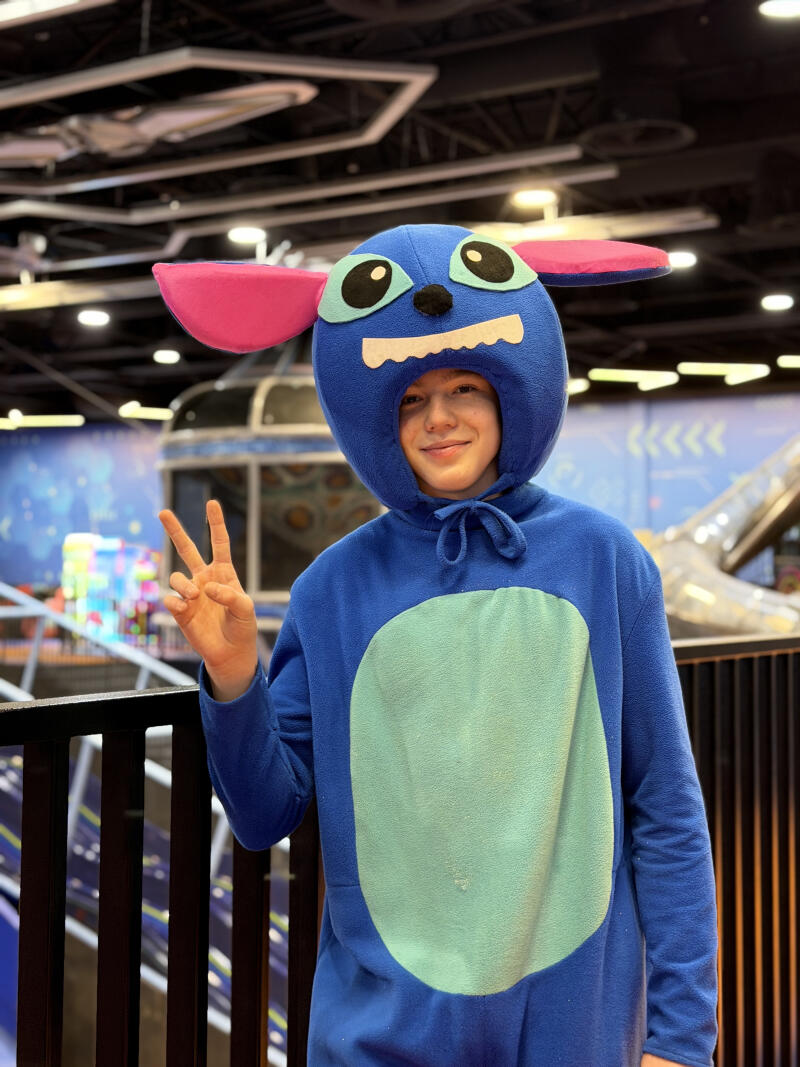 A person wearing a blue Stitch costume with pink ears and a light blue belly patch smiles and makes a peace sign. The background shows a blurred indoor entertainment area.
