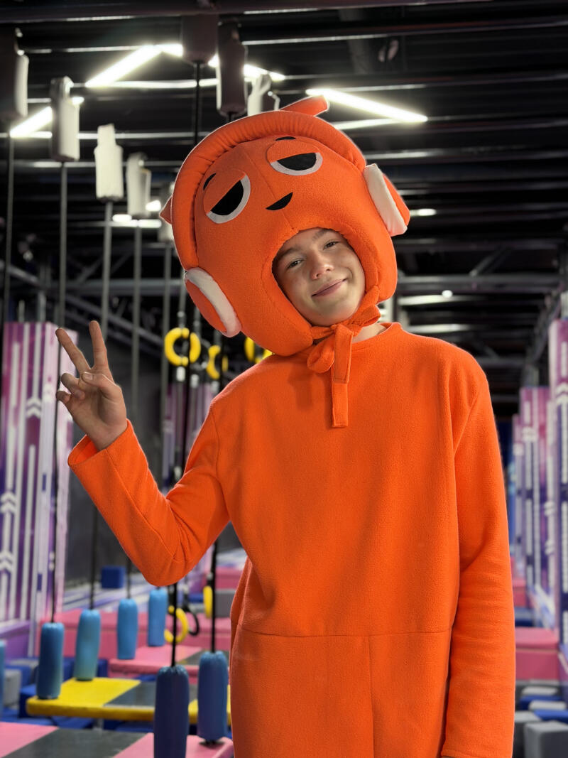 A young person wearing a vibrant orange full-body costume with a character head featuring large headphones, dark eyes, and a smile, makes a peace sign with their hand in an indoor play area.