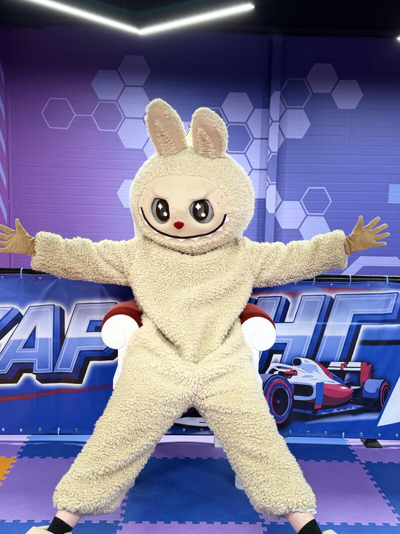 A person in a fluffy, light brown bunny mascot costume with large eyes and a wide smile stands with outstretched arms, leaning on a red and white chair. The background features purple and blue walls with hexagonal patterns and a banner displaying "AF HT" text and a racing car graphic, while the floor consists of interlocking purple and blue mats.