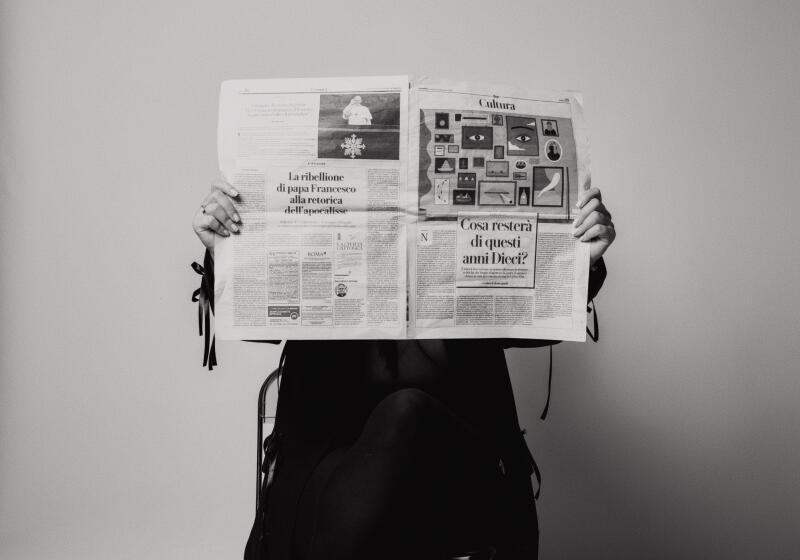 A black and white photo shows a person holding an open newspaper that covers their face, revealing only their hands and lower body. Visible headlines include 'La ribellione di papa Francesco alla retorica dell'apocalisse' and 'Cosa resterà di questi anni Dieci?', with a 'Cultura' section featuring illustrations of eyes.
