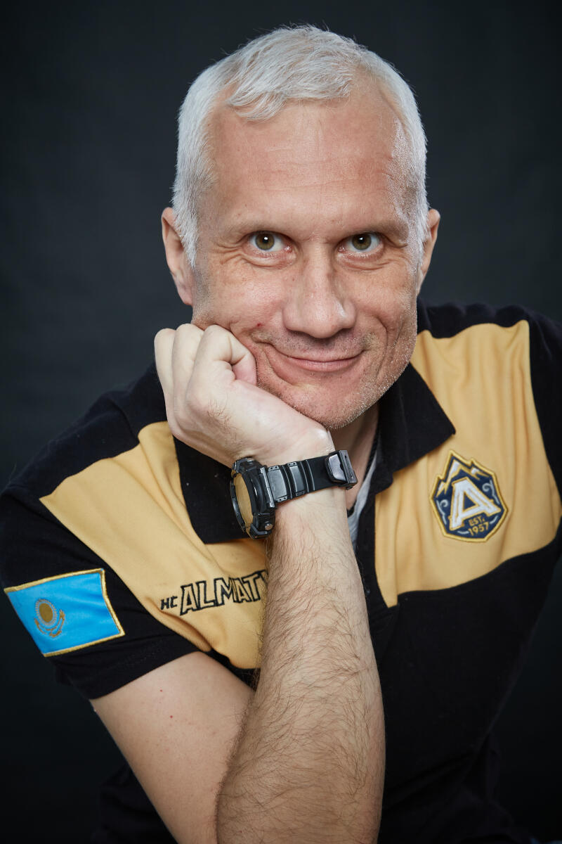 A smiling man with gray hair and green eyes rests his chin on his hand, wearing a black and gold polo shirt with a black digital watch. The shirt features a Kazakhstan flag patch and "HC ALMATY" text on the sleeve, and an emblem with "EST. 1957" on the chest.