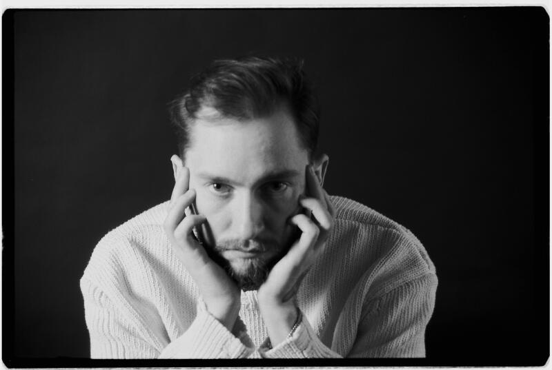A black and white portrait of a bearded man wearing a light-colored knitted sweater, looking intently at the camera with his hands placed by his temples.