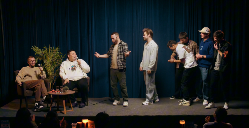 Two men are seated on the left side of a stage, both holding microphones and smiling, while six other men stand to their right. A dark blue curtain forms the backdrop, and an audience is visible in the foreground.