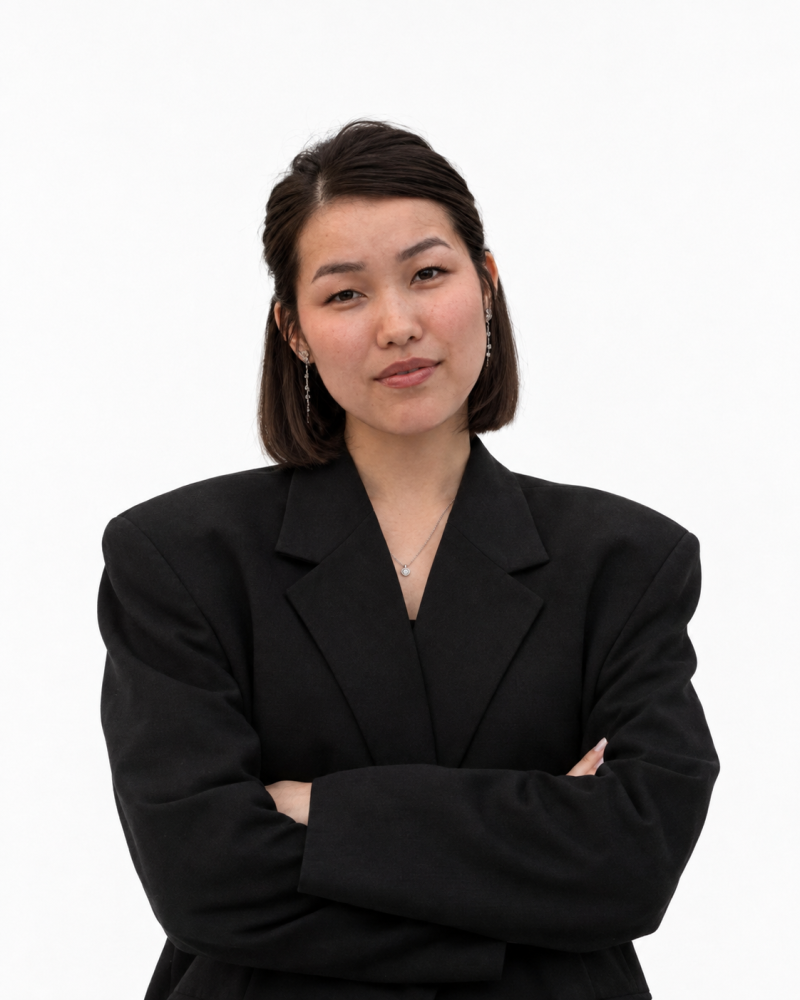 A young Asian woman with short dark hair and a black blazer stands with her arms crossed, looking at the camera against a white background. She wears a silver necklace and dangle earrings.