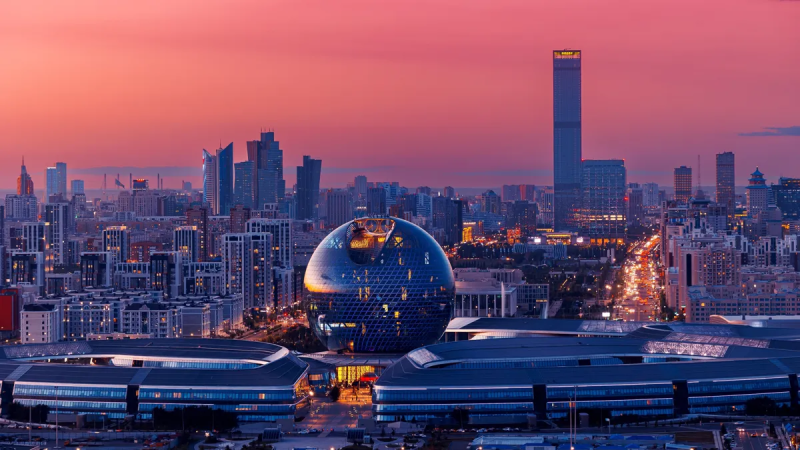 An aerial view of a modern city at dusk, featuring a large spherical building with a patterned glass facade, surrounded by other skyscrapers and residential buildings, a long street with illuminated car lights, all under a vibrant pink and orange sky.