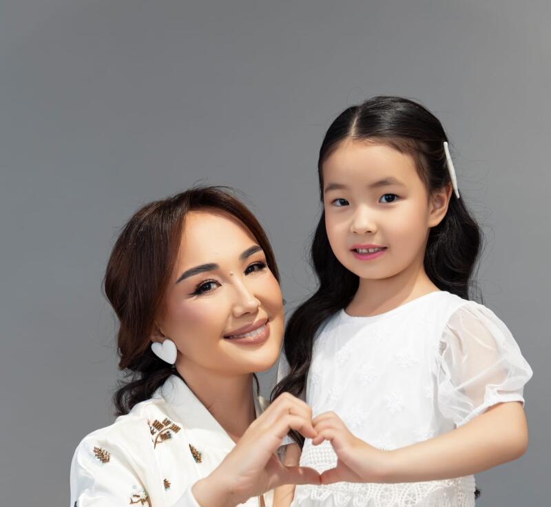 A smiling woman and a young girl make a heart shape with their hands, both looking at the camera against a gray background. The woman wears a white embroidered top and heart earrings, while the girl wears a white dress.