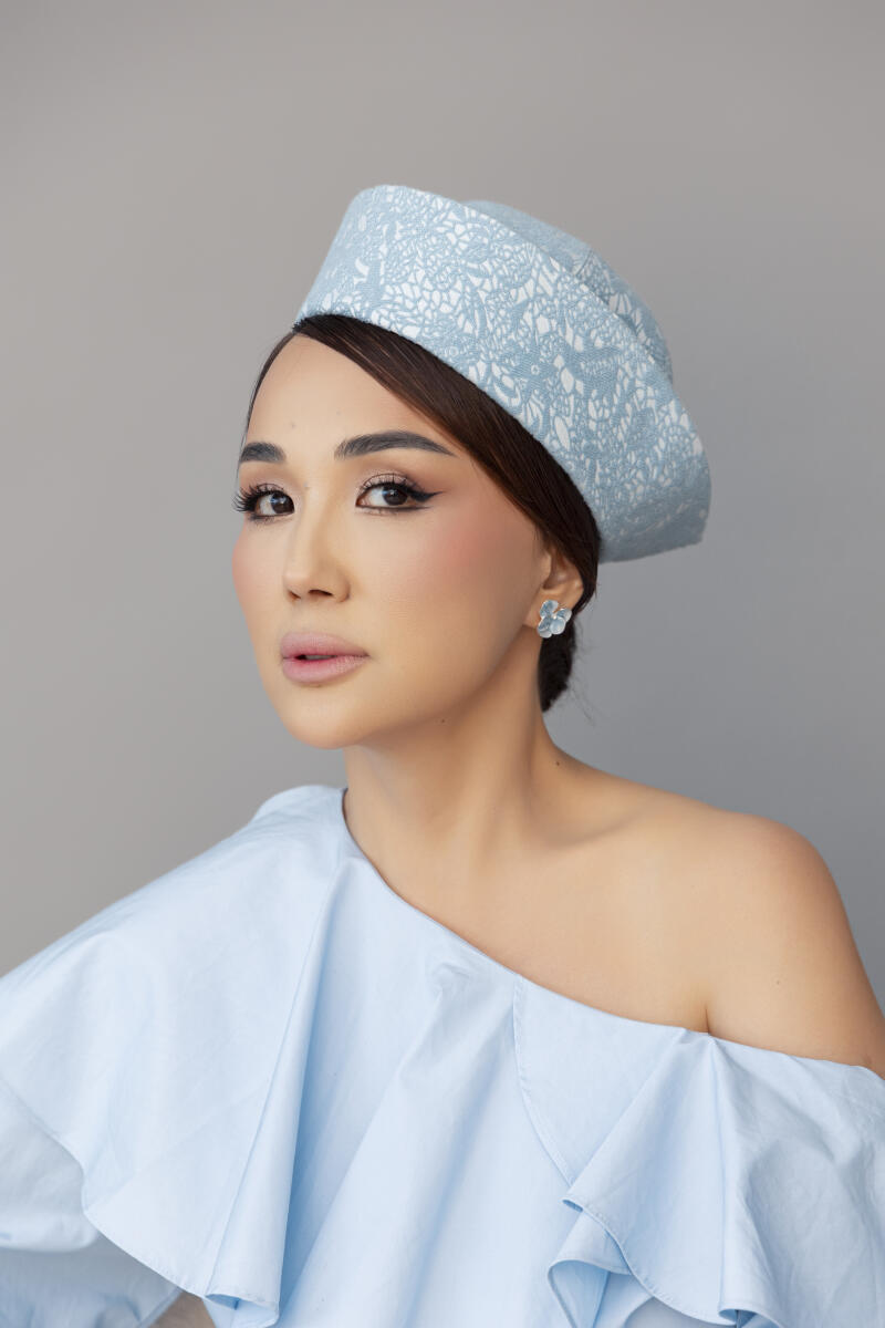 A woman wearing a light blue hat with a white lace pattern and a matching off-the-shoulder top, adorned with a light blue flower earring and looking directly at the camera.