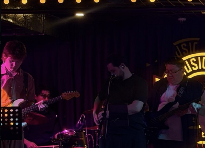 A band performs on a dark stage illuminated by purple lights, with four members visible playing electric guitars, bass, and drums, and a neon "MUSIC" sign in the background.