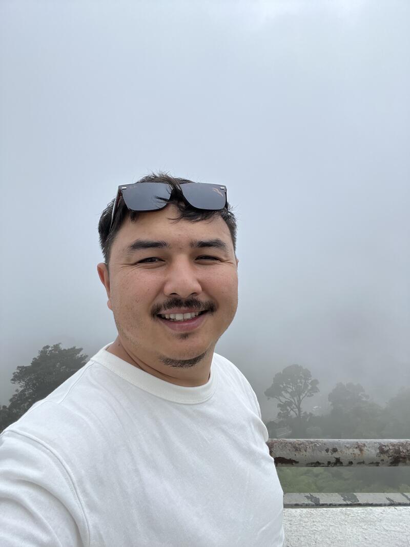 A smiling man in a white t-shirt with sunglasses on his head takes a selfie against a foggy background with faint trees and a rusty railing visible below.