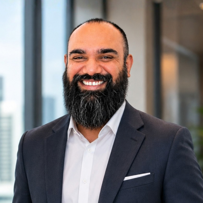 A smiling man with a dark, full beard and a shaved head wears a dark suit and a white shirt with a pocket square, against a blurred background of large windows.