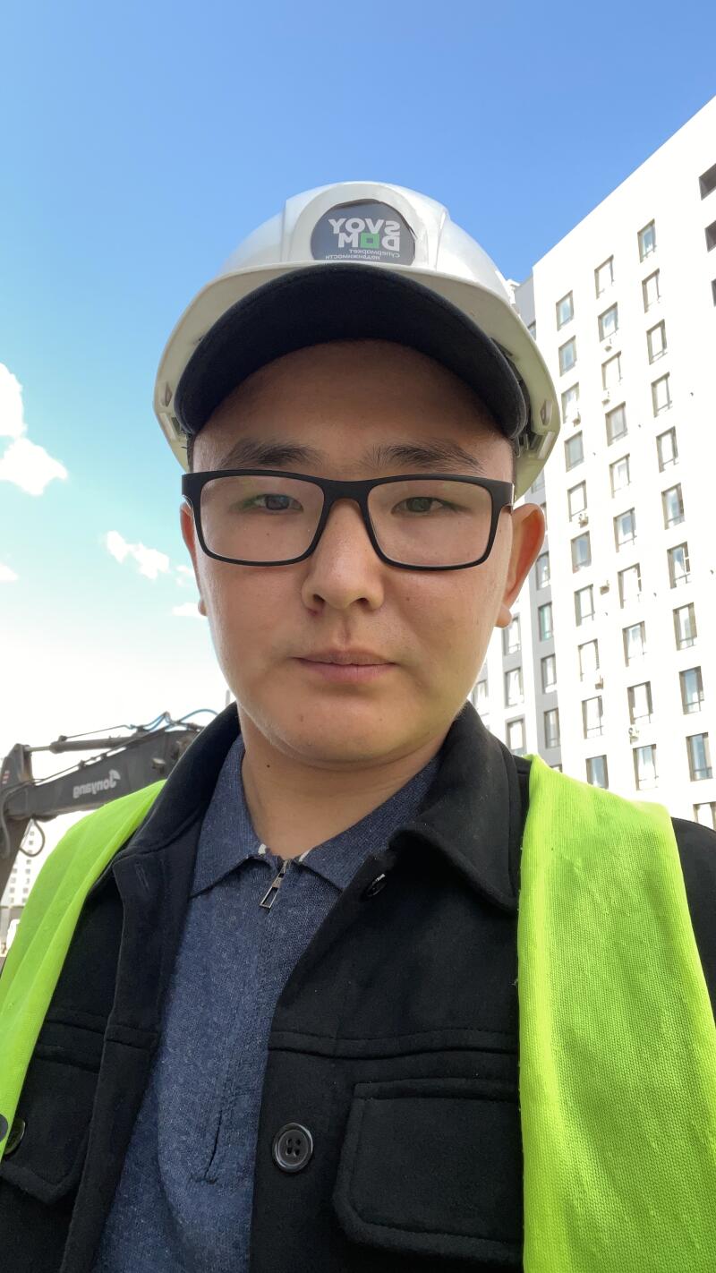 A man wearing glasses, a white hard hat with a reversed "YOVS MД" logo, a black jacket, and a neon yellow safety vest. An excavator with reversed "ensynot" text and a white multi-story building are in the background under a blue sky.