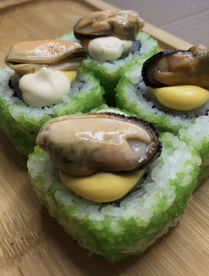 Close-up of sushi rolls topped with mussels, some featuring white sauce and others yellow sauce, all covered in green tobiko and served on a wooden surface.