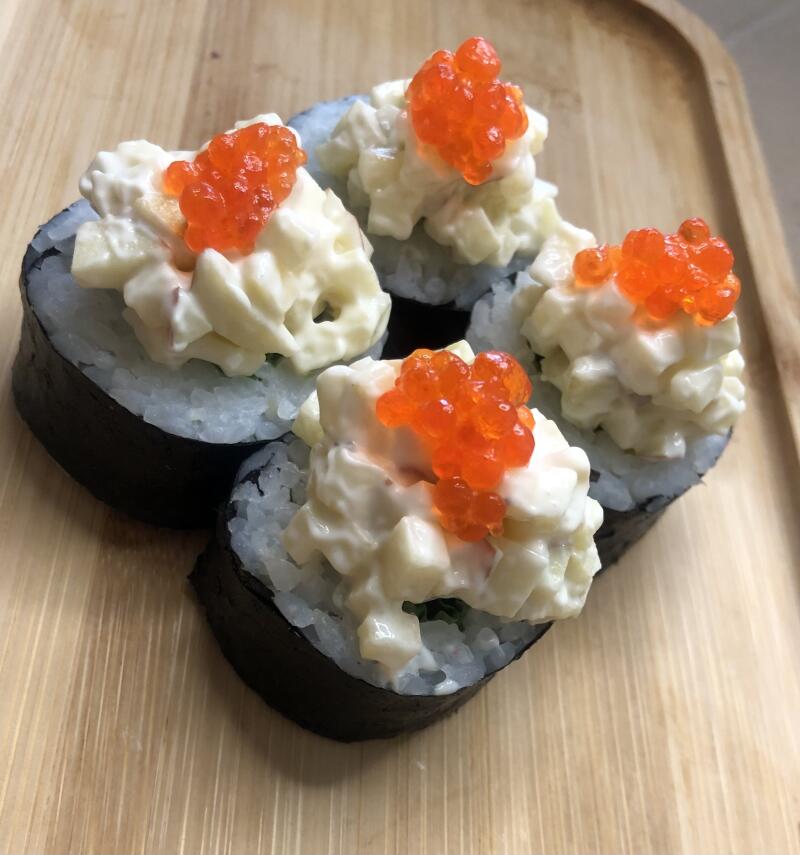 Four maki sushi rolls with rice and nori, topped with a creamy, diced white mixture and garnished with bright orange caviar, served on a light wooden tray.