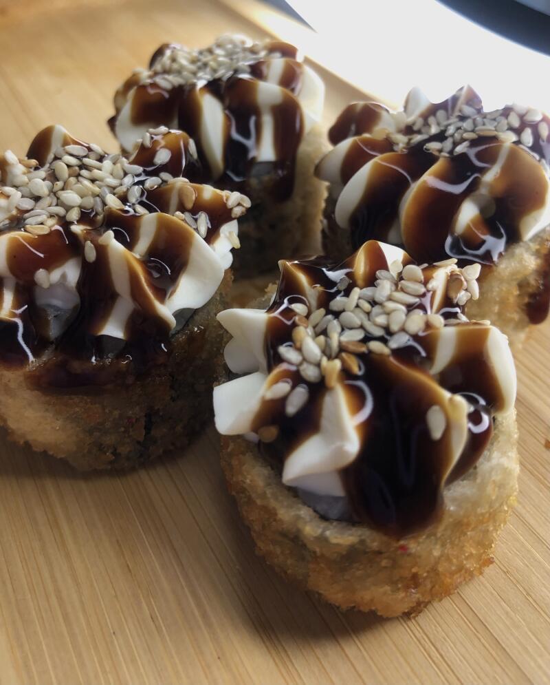 Close-up of several deep-fried sushi rolls topped with white cream, dark sauce, and sesame seeds, arranged on a light brown wooden surface.