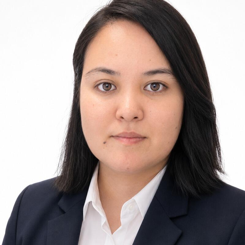 A headshot of a woman with dark hair and brown eyes, wearing a dark blue suit jacket over a white collared shirt, against a plain white background.