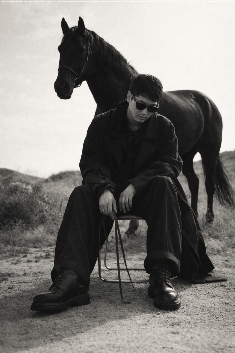A black and white photo shows a man sitting on a chair, wearing a dark coat, wide-leg pants, boots, and sunglasses, holding a strap, with a dark horse standing behind him in an outdoor setting.