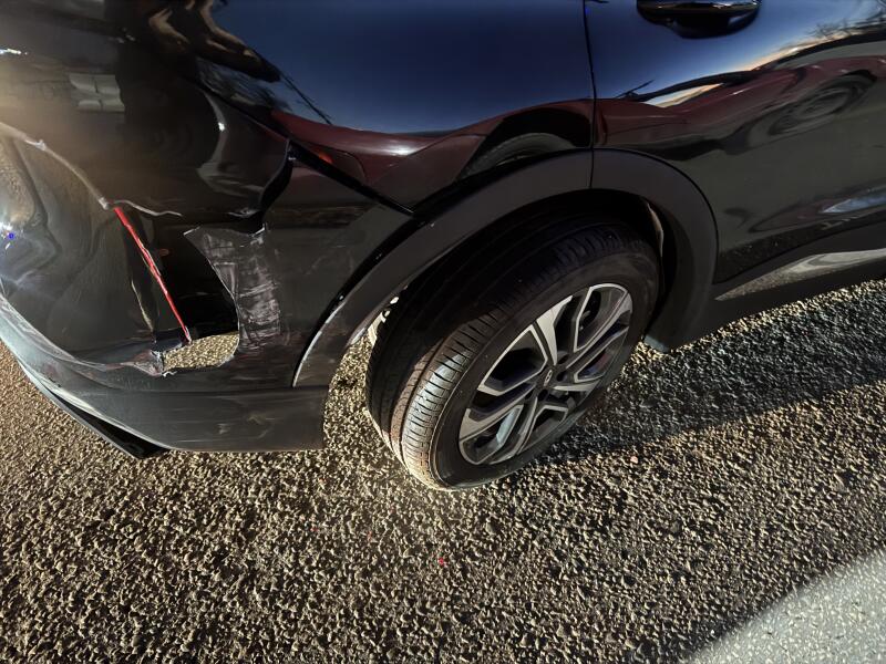 A close-up view of the rear left side of a black car showing significant damage to the bumper and fender, with a piece of the fender detached and visible scratches. The car is parked on a gravel surface.