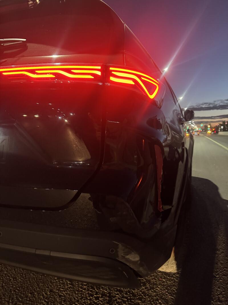 Rear view of a dark car at dusk, showing bright red taillights with yellow LED accents. The lower right side of the bumper has visible damage, including a dent and torn red material, with blurred city lights in the background.