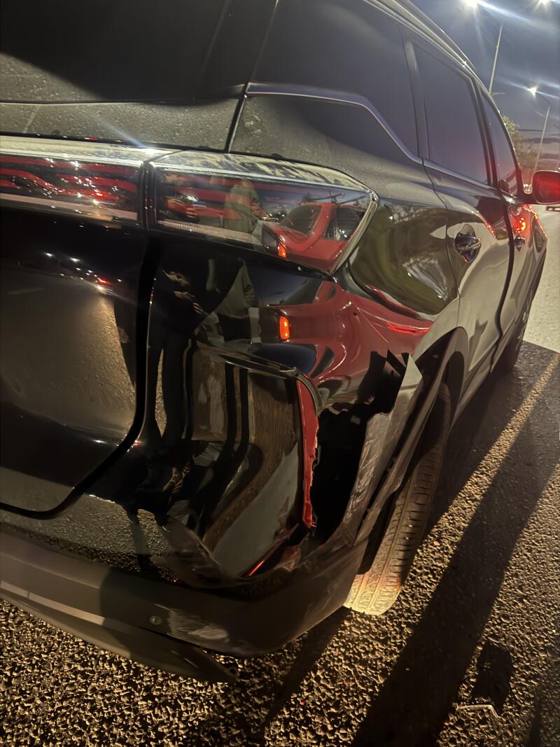 The rear-left side of a black car is shown at night, featuring a dent on the fender with red marks and reflections of a person holding a phone and streetlights.
