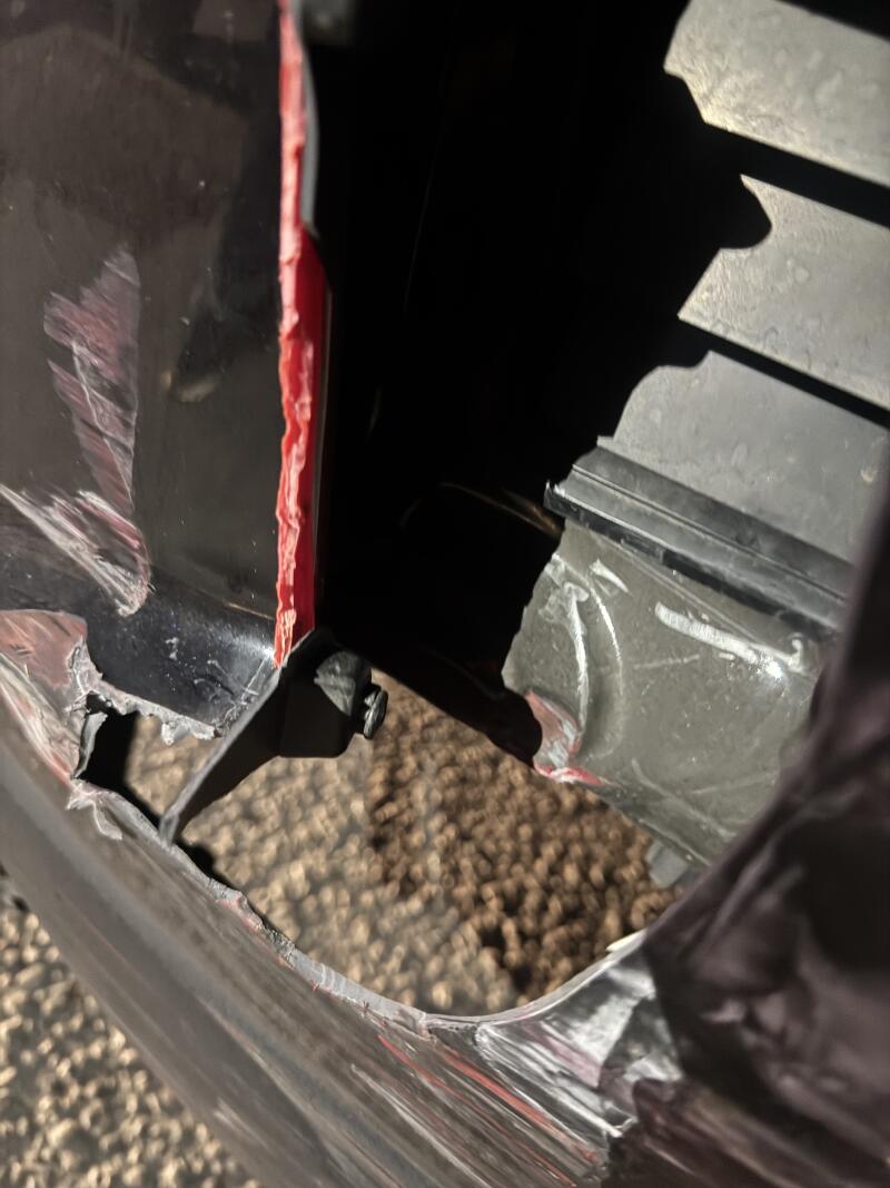 A close-up of a damaged black vehicle bumper or panel, showing a tear exposing red paint and a visible bolt. The surface has scratches and peeling clear film, with a textured brown ground visible through the opening.