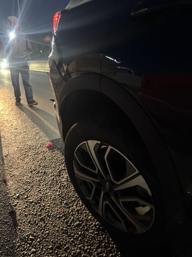 A dark car is parked on an asphalt road at night, with its taillight glowing red and a person holding a flashlight in the background, with red debris on the ground.