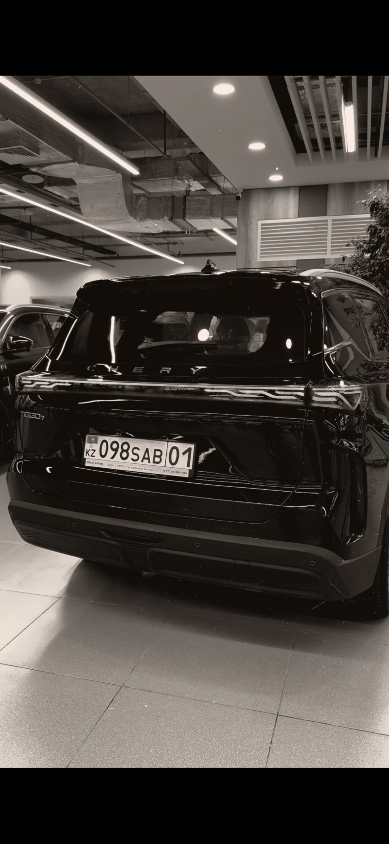 A monochromatic rear view of a black Chery Tiggo 7 SUV, displaying the Kazakhstan license plate KZ 098SAB 01 and dealer information, parked indoors under industrial-style lighting.