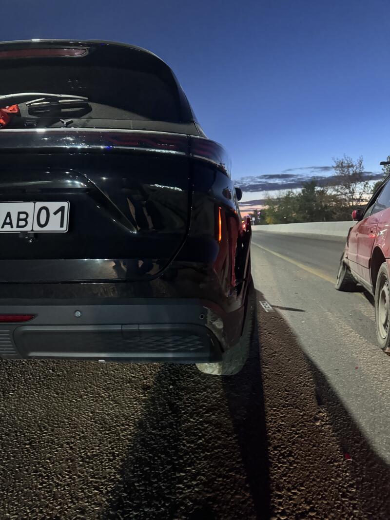 The rear view of a black car with license plate 'AB 01' parked on the side of a road, next to a damaged red car, under a dusk or dawn sky.