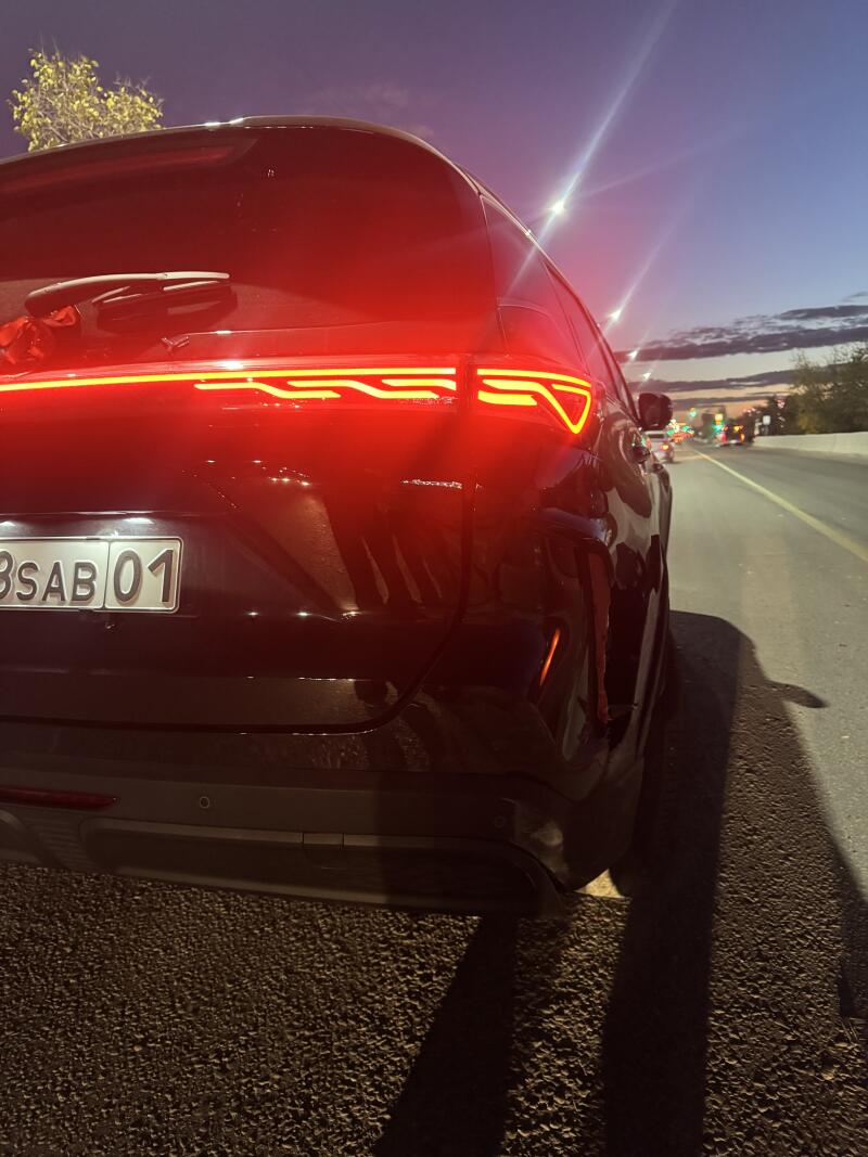 A black car is seen from the rear, parked on the side of a road at dusk or night. Its red taillights are illuminated with a distinctive horizontal line and a 'V' shape, and the license plate reads 'BSAB 01'.