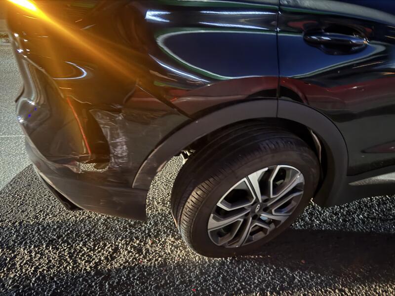 The rear side of a black car is shown with visible damage, including scratches and a dent on the bumper and fender, reflecting a bright orange light.