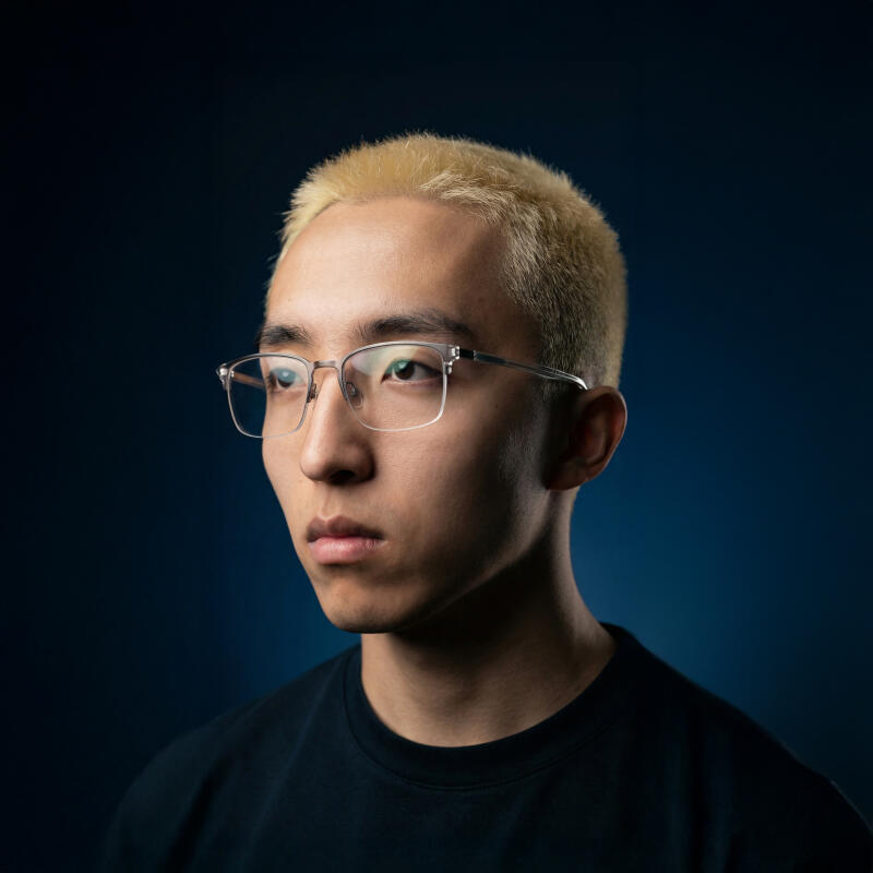 A young man with light blonde hair and clear-framed glasses looks to the left against a dark blue background.