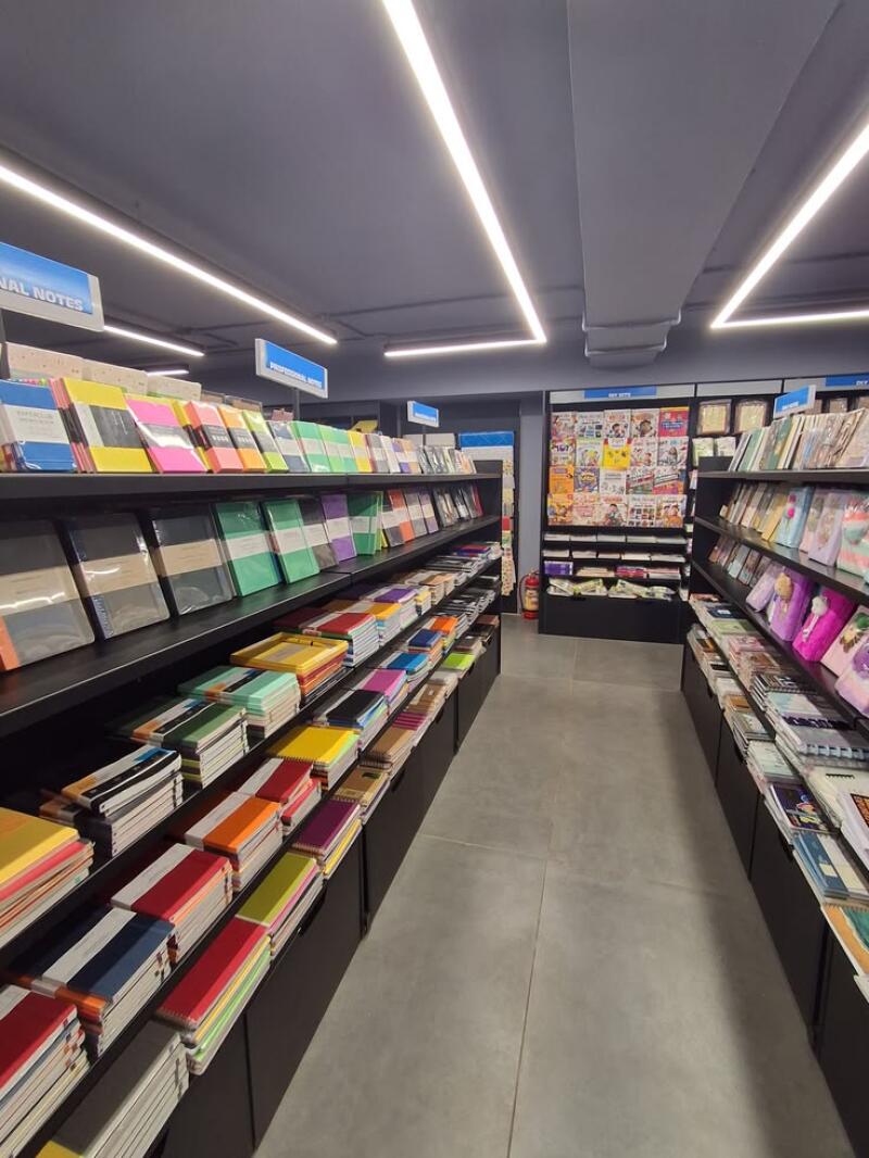 An aisle in a stationery store with shelves packed with colorful notebooks and journals, illuminated by linear ceiling lights. Signs visible include "PERSONAL NOTES", "PROFESSIONAL NOTES", and "DIY KITS".