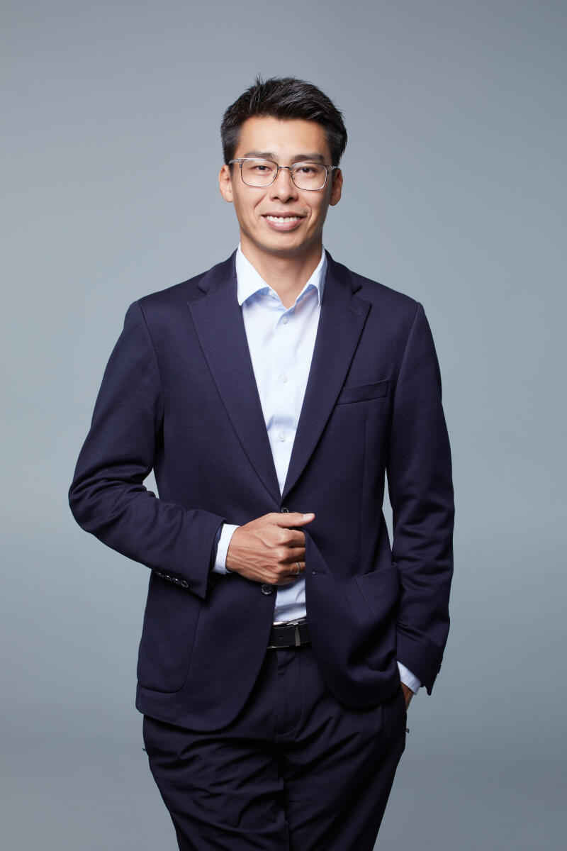 A smiling man wearing glasses, a dark blue suit jacket, and a light blue shirt poses against a grey background. His right hand is on his jacket, and his left hand is in his pocket.