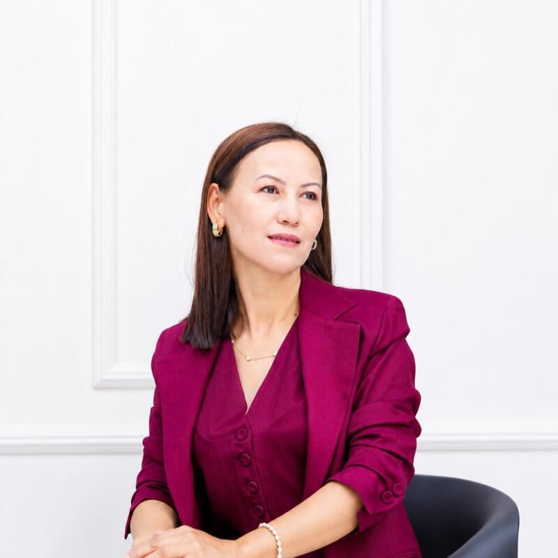 A woman with long dark hair, wearing a magenta suit, gold earrings, and a pearl bracelet, is seated and looking to the right against a white wall with decorative molding. Her hands are clasped in front of her.