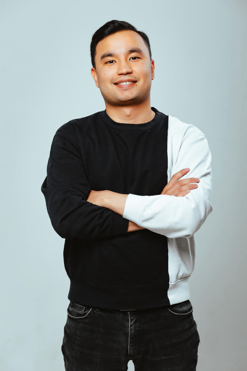 A smiling man with short dark hair is standing with his arms crossed, wearing a black and white color-block sweatshirt and dark jeans, against a light gray background.