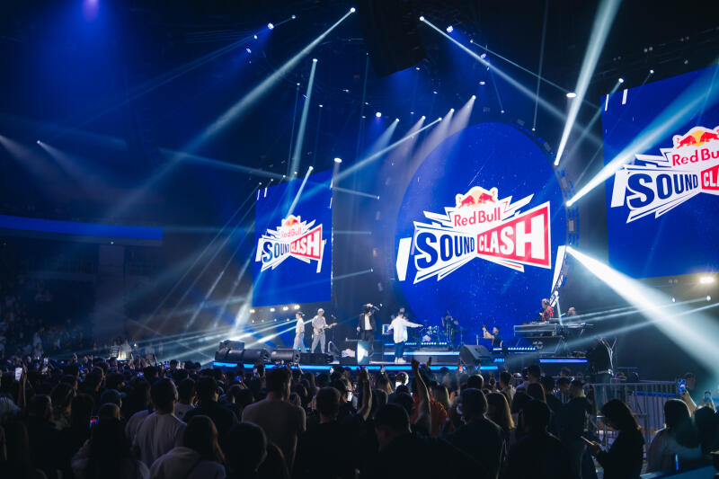 A concert stage with a band performing in front of a large crowd, featuring Red Bull SoundClash branding on the screens.
