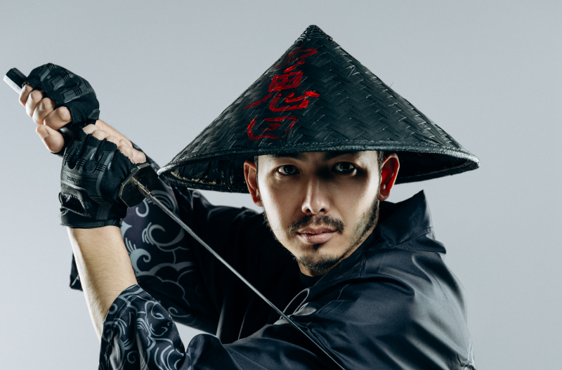 A man in a conical hat with red symbols and black gloves holds a katana.