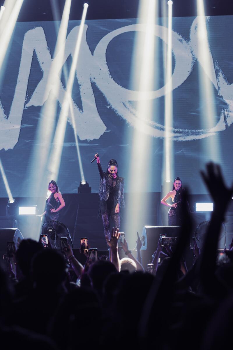 Three women are on stage during a concert, illuminated by bright spotlights, with a backdrop displaying a large white logo and a crowd of people holding up their phones to record the performance.