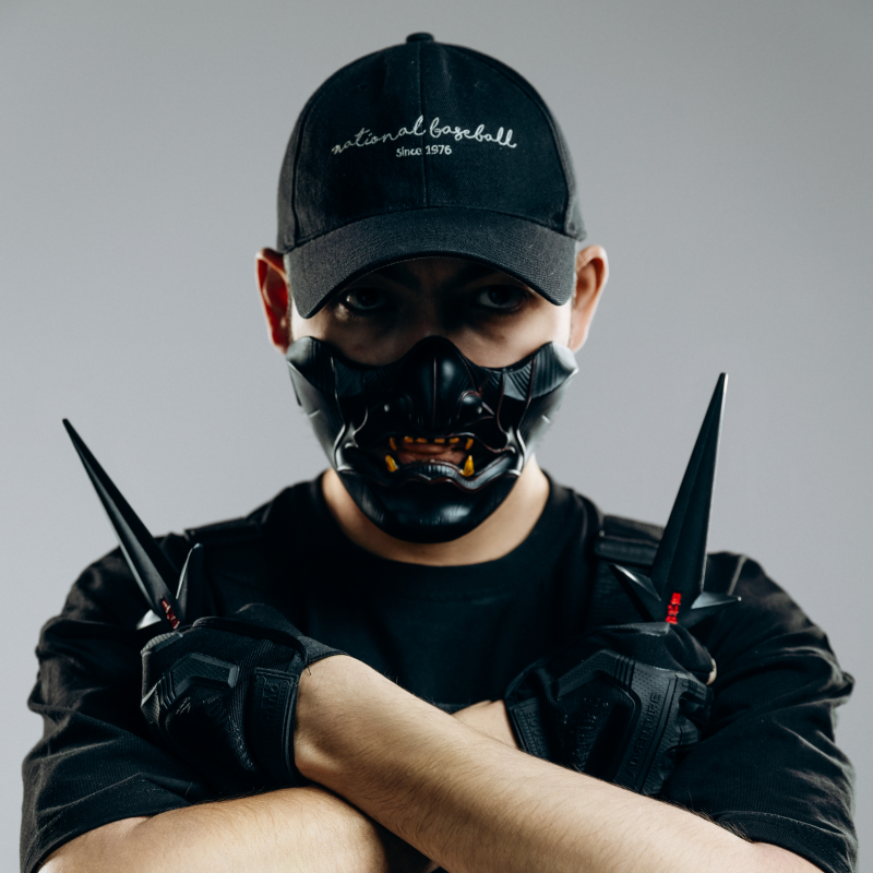 A person wearing a black "national baseball since 1976" cap and a demon-like mask is holding two sharp weapons while crossing their arms.