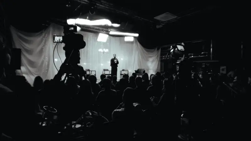 A black and white photo shows a speaker on a stage addressing a dimly lit audience, with a camera in the foreground.