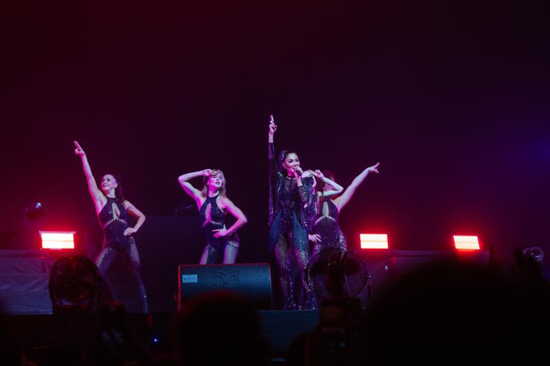 A singer performs on stage with three backup dancers, all wearing sparkly dark outfits, under bright stage lights.