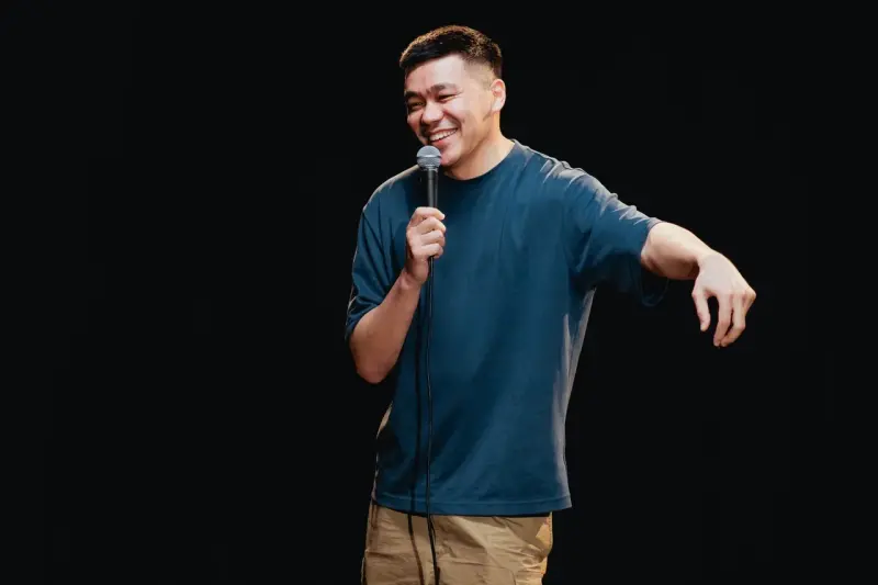 A man with short dark hair, wearing a blue t-shirt and khaki shorts, is smiling and holding a microphone in his right hand against a black background.