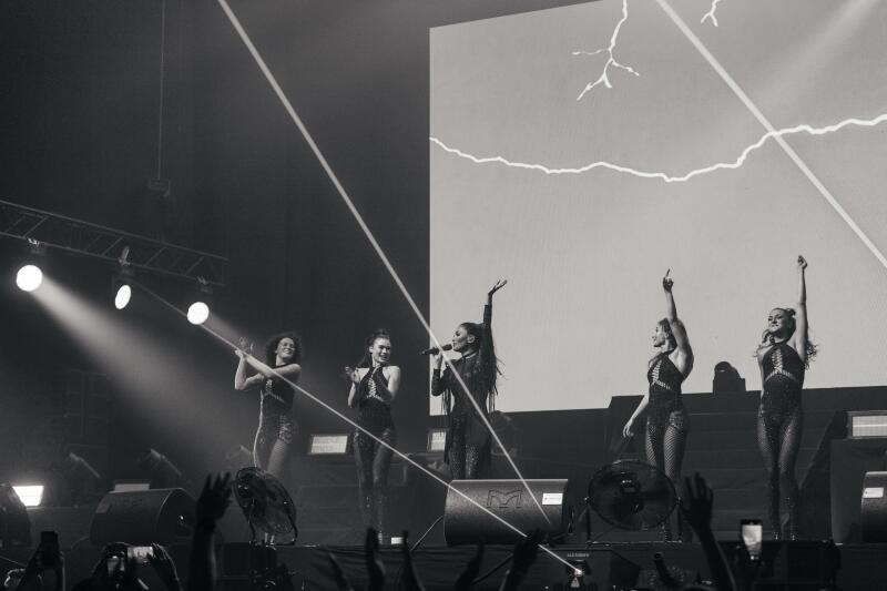 A black and white photo captures a musical performance with five women on stage in dark, sparkly outfits, backed by a screen displaying a lightning bolt graphic, and the audience is visible in the foreground, with many holding up their phones.