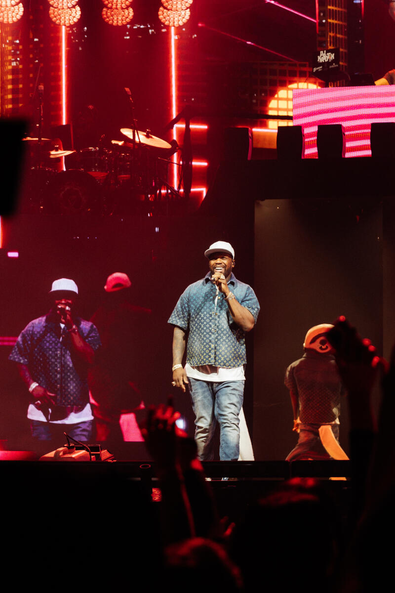 50 Cent is performing on stage in a blue shirt and jeans with a white baseball cap, holding a microphone while standing in front of a large screen with red lighting and the words "DJ Khaled" visible in the upper right corner.