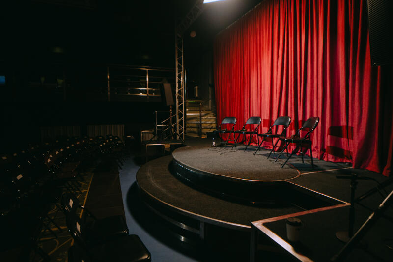 The stage is set with a red curtain backdrop, a tiered platform, and four black folding chairs lined up on the top tier. Rows of empty chairs fill the foreground, suggesting an auditorium.