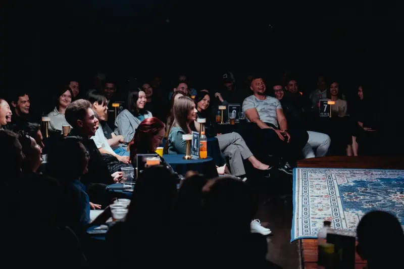A dimly lit room shows a crowd of people sitting at tables with drinks, many laughing, and a stage area with a patterned rug.