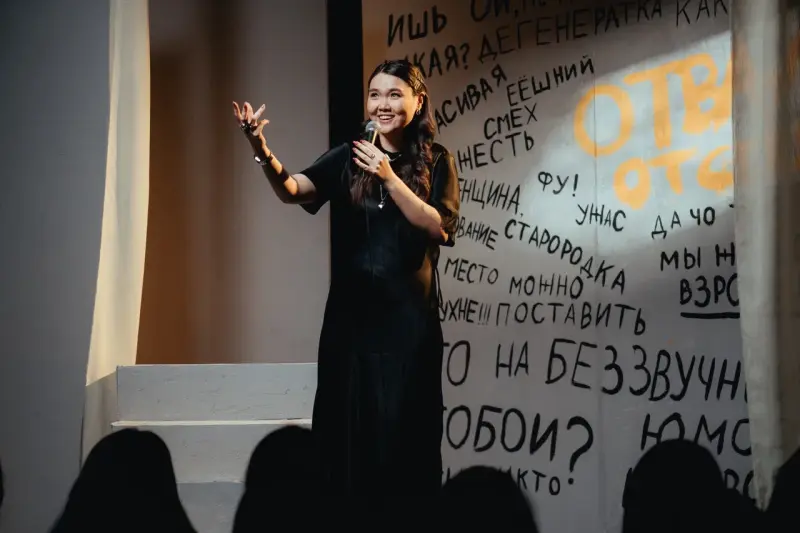 A woman in a black dress is on stage, holding a microphone and gesturing with her hand, in front of a wall covered in black and orange graffiti.
