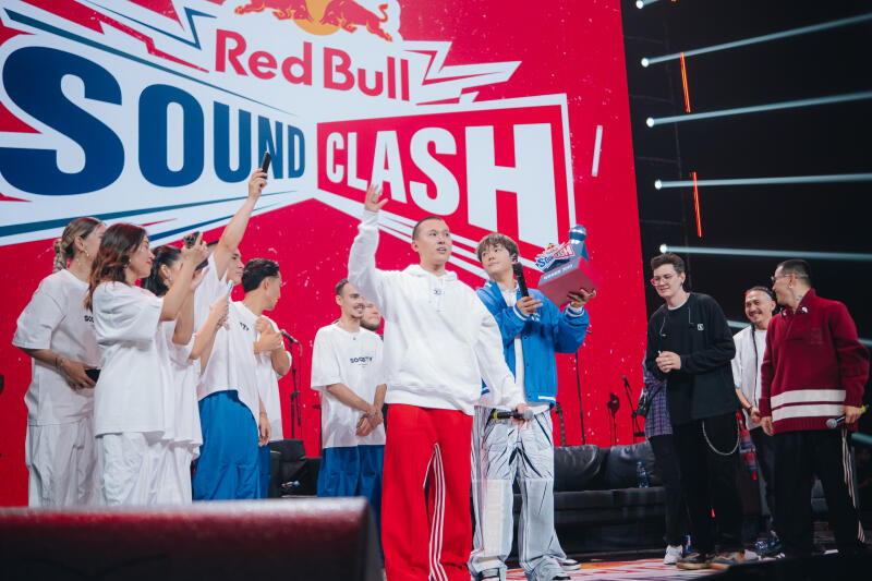 A group of people stand on a stage in front of a "Red Bull SOUND CLASH" backdrop, some are taking photos with their phones, while one man in a white hoodie and red pants raises his hand.