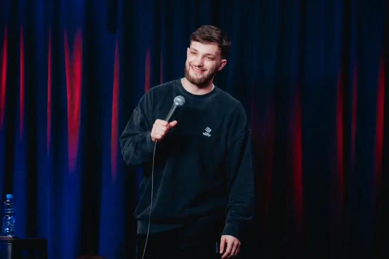 A man with a beard, wearing a black sweatshirt, holds a microphone and smiles in front of a blue and red curtain.