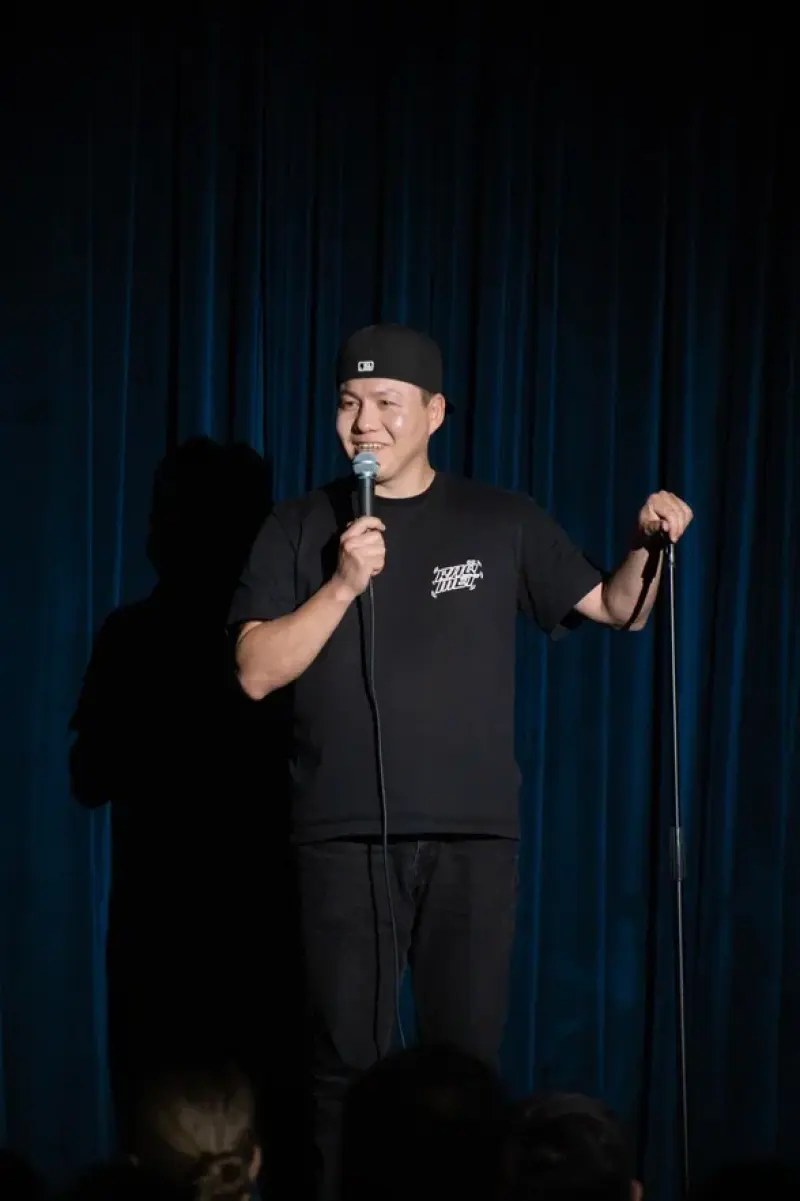 A man in a black t-shirt and baseball cap is on stage, holding a microphone in one hand and the mic stand in the other, with a blue curtain in the background.