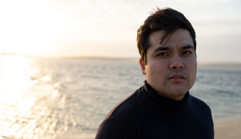 Мужчина в черной водолазке позирует на фоне водоема, отражающего солнечный свет.